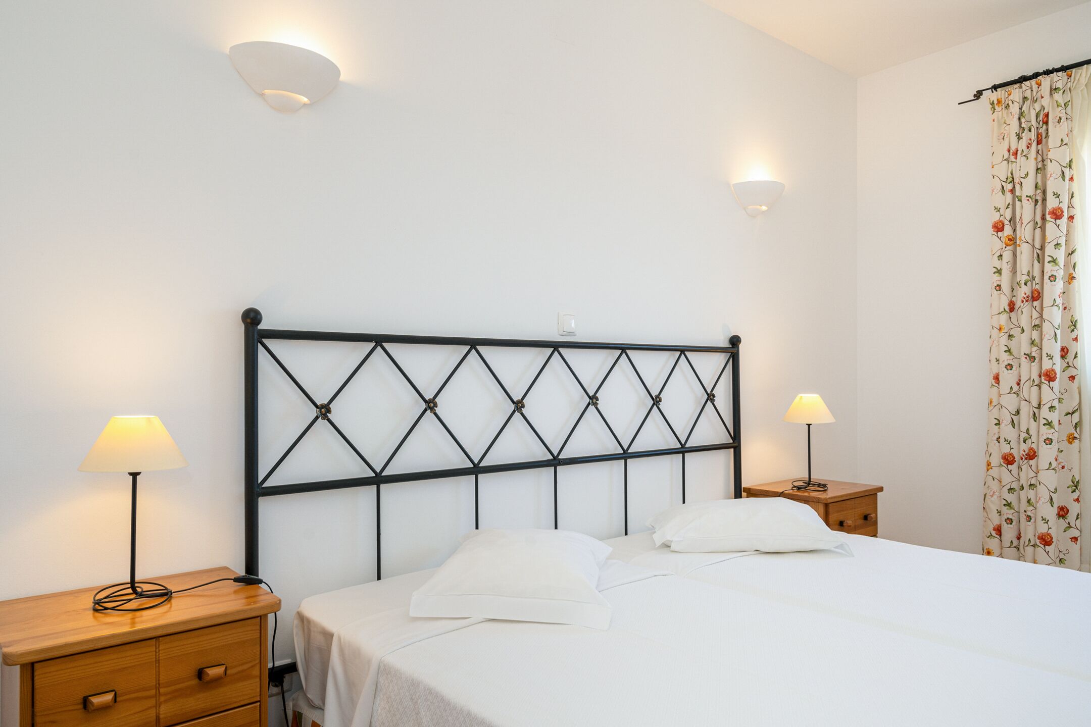 A neatly made bed with a headboard, two bedside tables, and a floral curtain.