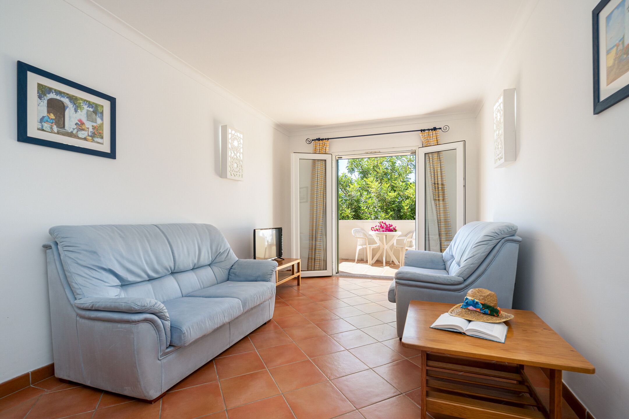 A living room with a sofa, a chair, a wooden table, and a balcony with a view of greenery.
