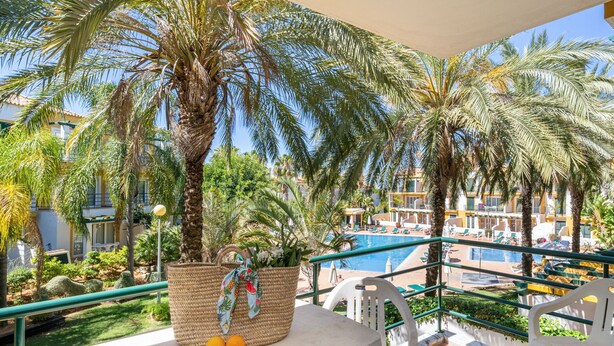 A balcony with a view of a pool, palm trees, and a building with multiple floors. - Quinta do Morgado - Monte da Eira (Tavira)