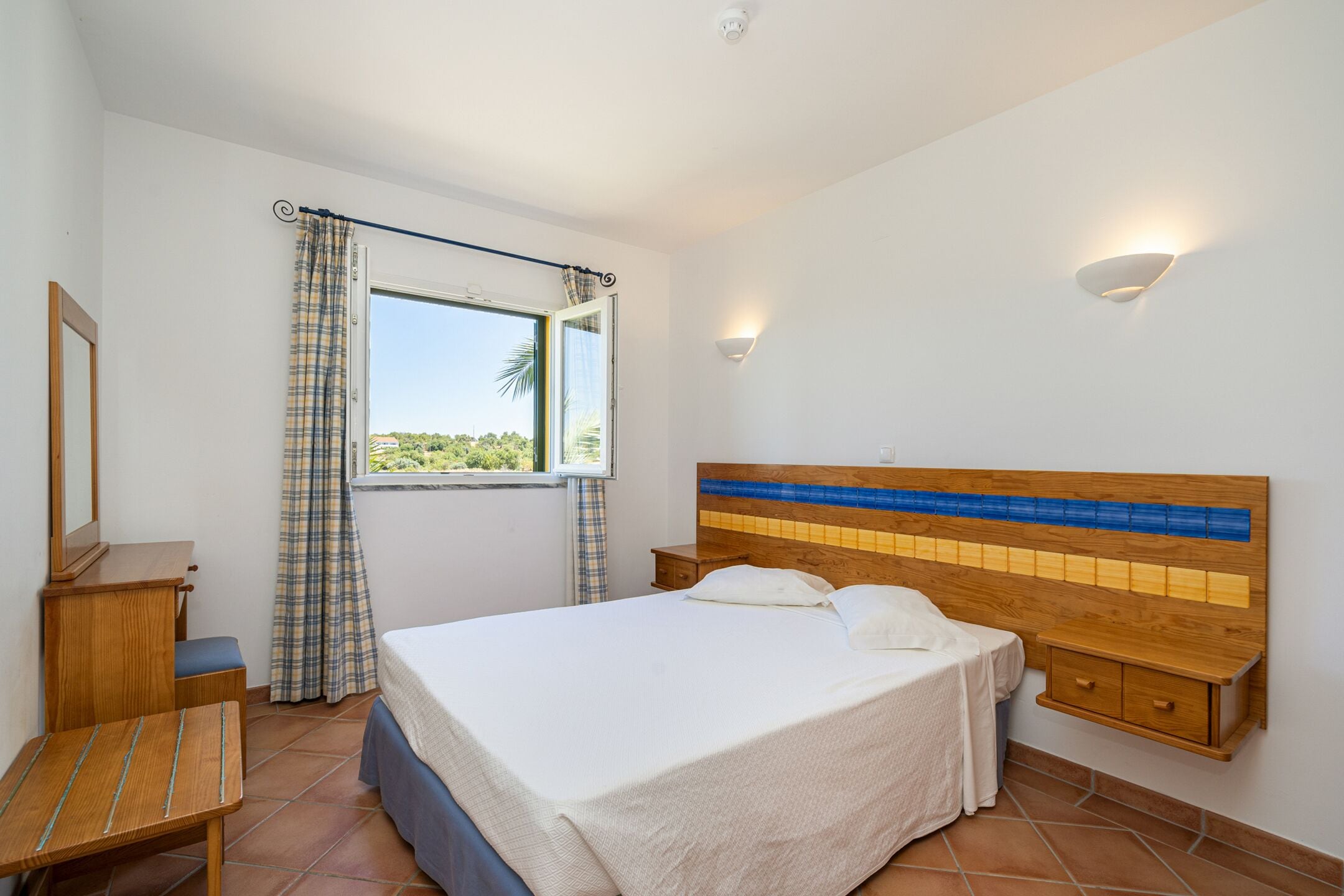 Une chambre à coucher comprenant un lit, une table de chevet, une tête de lit en bois, une fenêtre avec des rideaux et un luminaire fixé au mur.