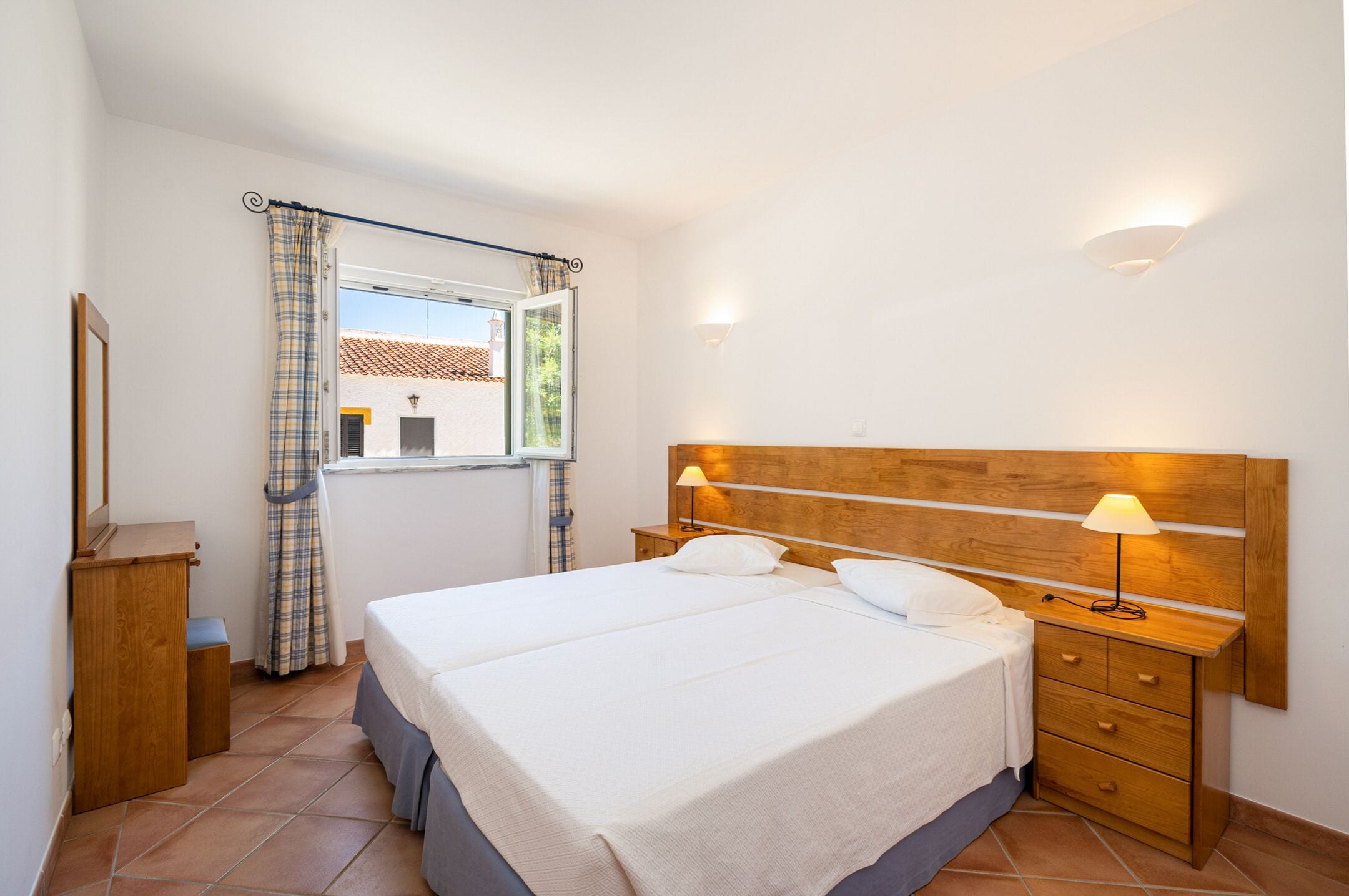 A bedroom with a large bed, two bedside tables, a nightstand, a window with curtains, and a wall-mounted lamp.