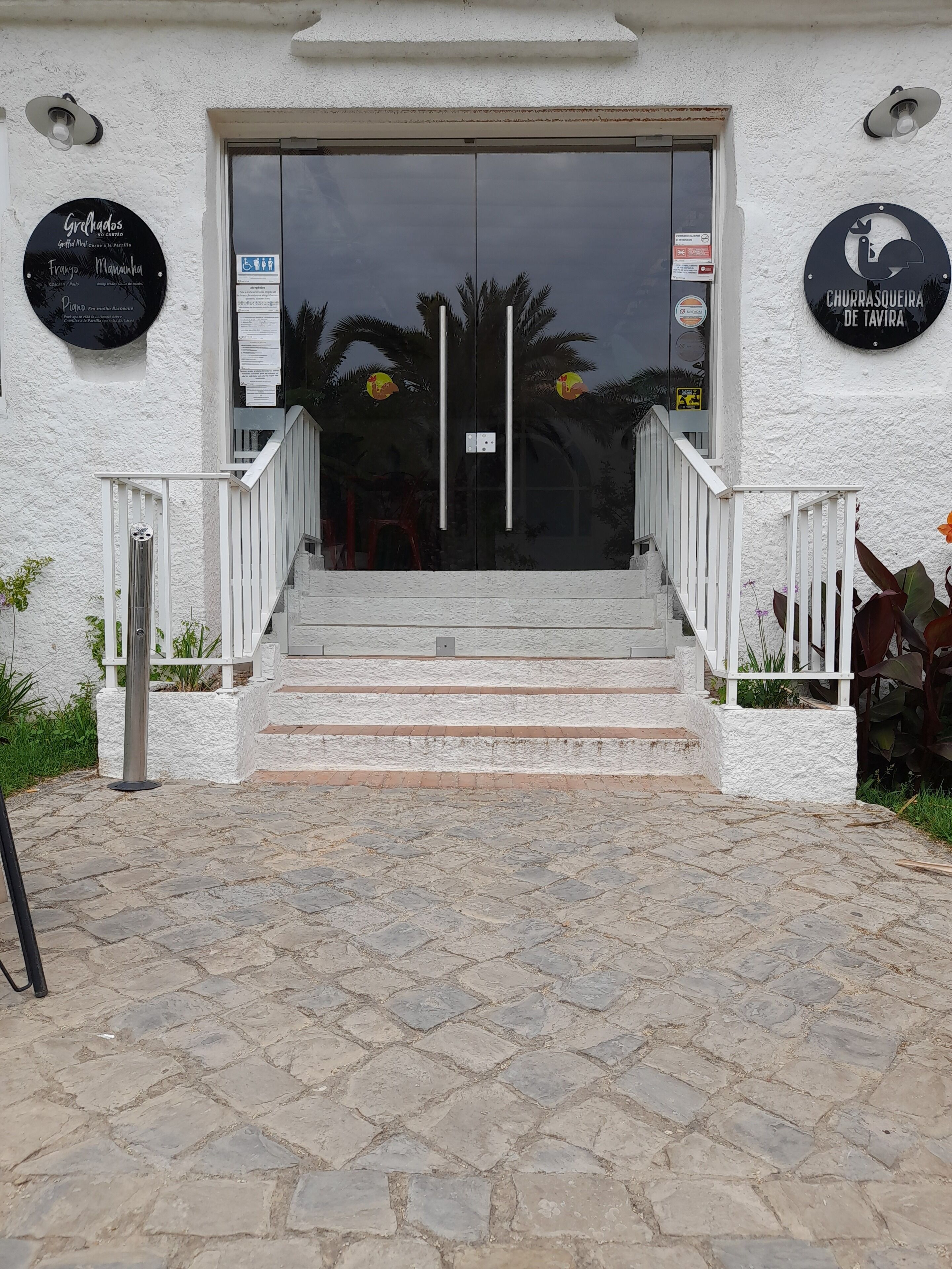La entrada de un edificio con un camino adoquinado que lleva a un conjunto de escalones. Hay dos carteles a ambos lados de la entrada, uno sobre la puerta y otro en la pared a la derecha.