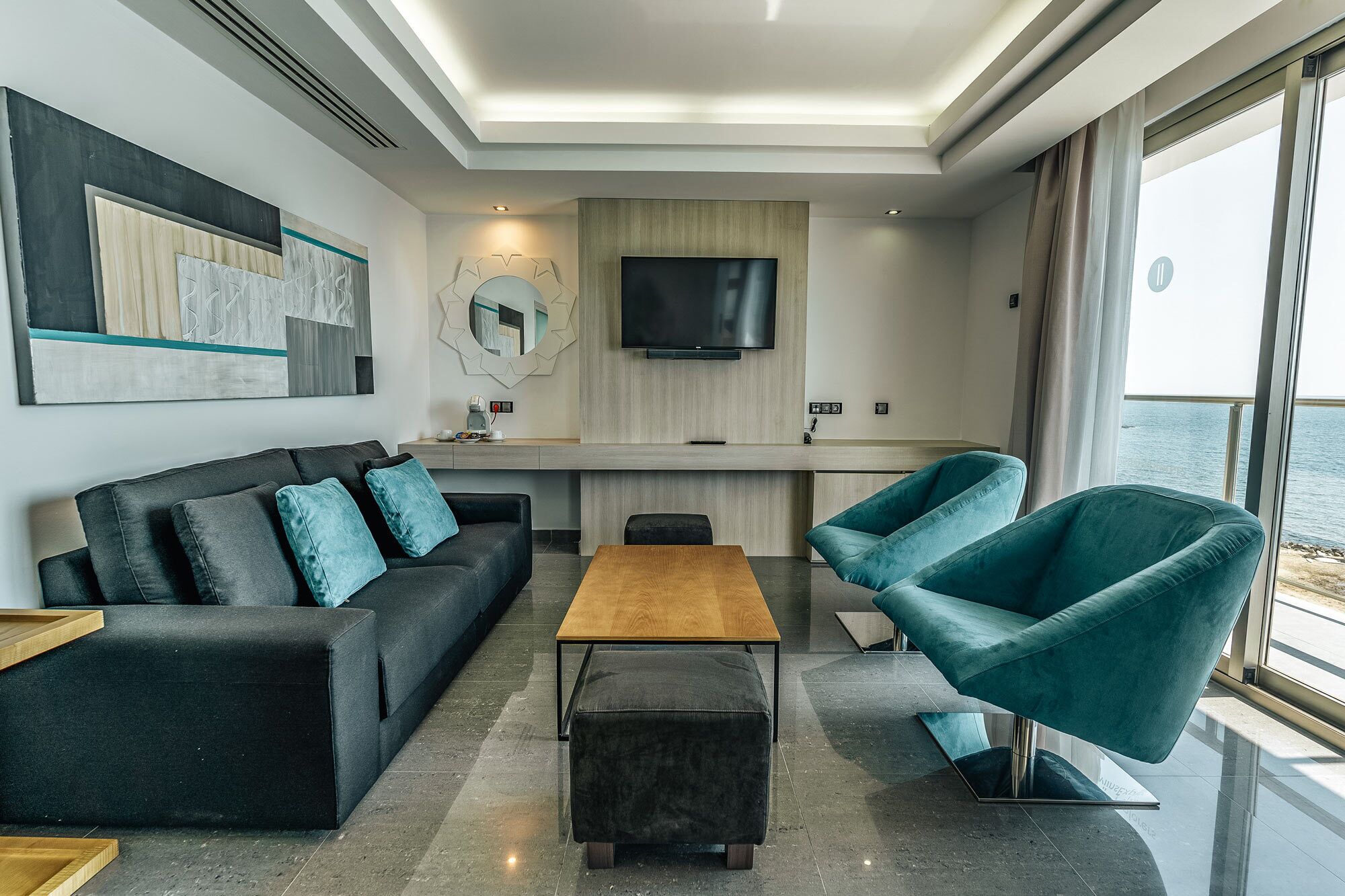 A modern living room with a sofa, ottoman, and teal armchairs. A wooden coffee table is in the center. A flat-screen TV is mounted on the wall above a wooden console table.