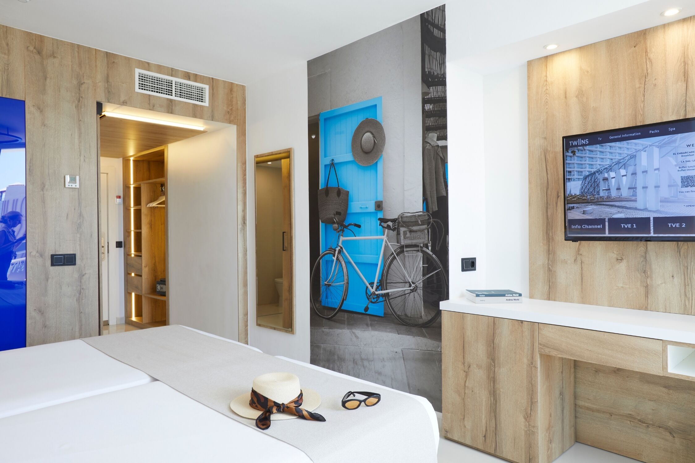 A modern hotel room with a bed, a TV, a wardrobe, and a wall with a bicycle and a blue door.