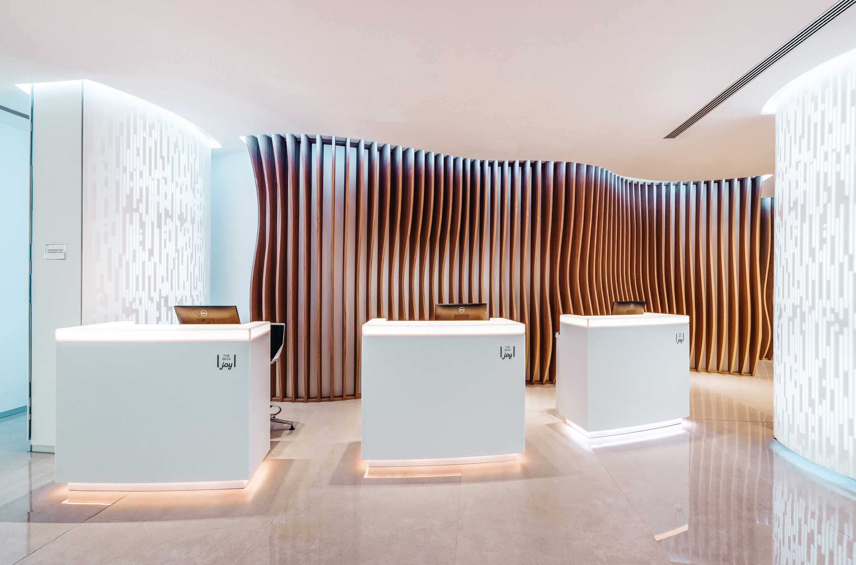 A reception area with three white reception desks, each with a laptop and a small table. The walls have a modern design with vertical wooden slats.