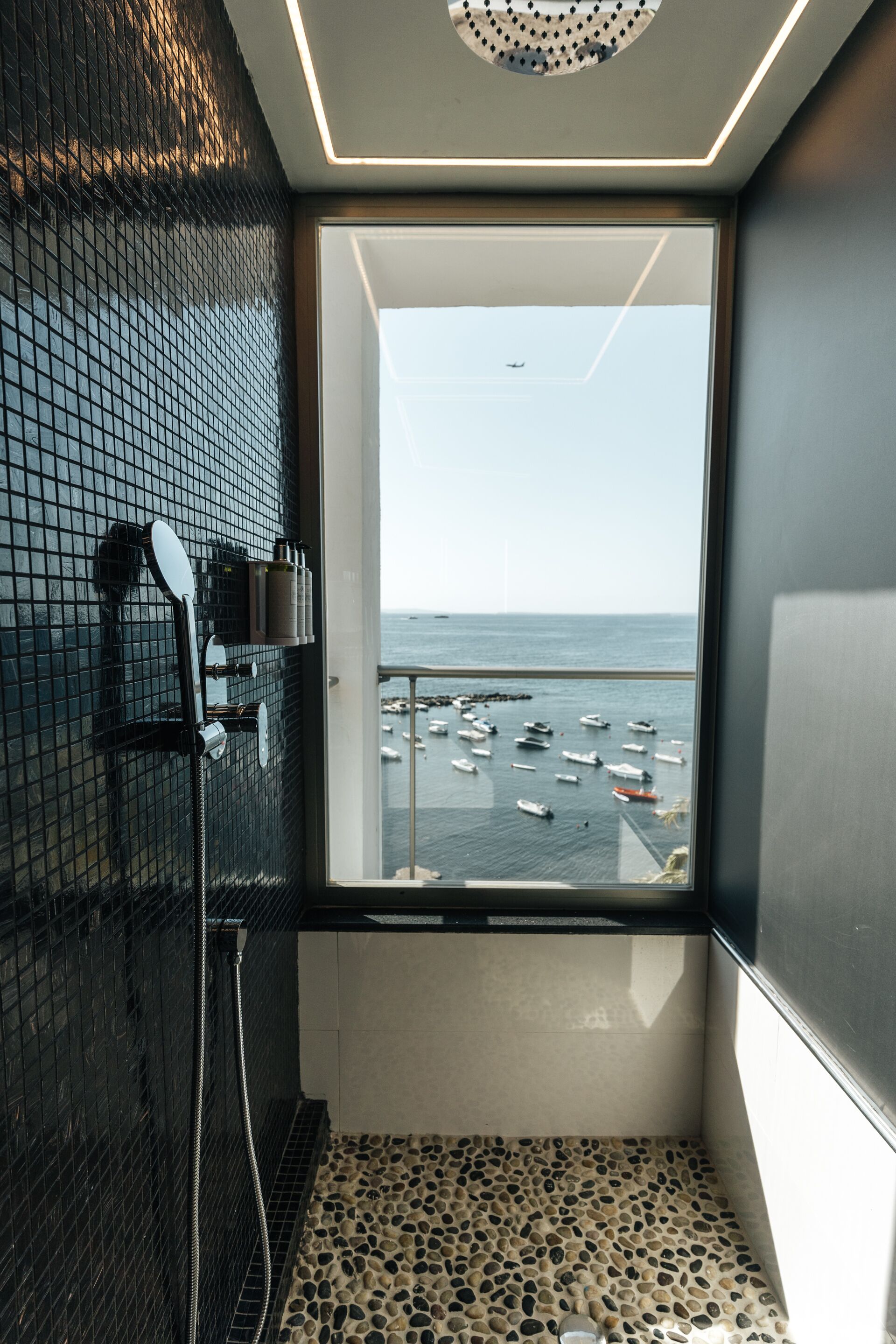 A bathroom with a tiled floor, a shower, and a window with a view of boats on the water.