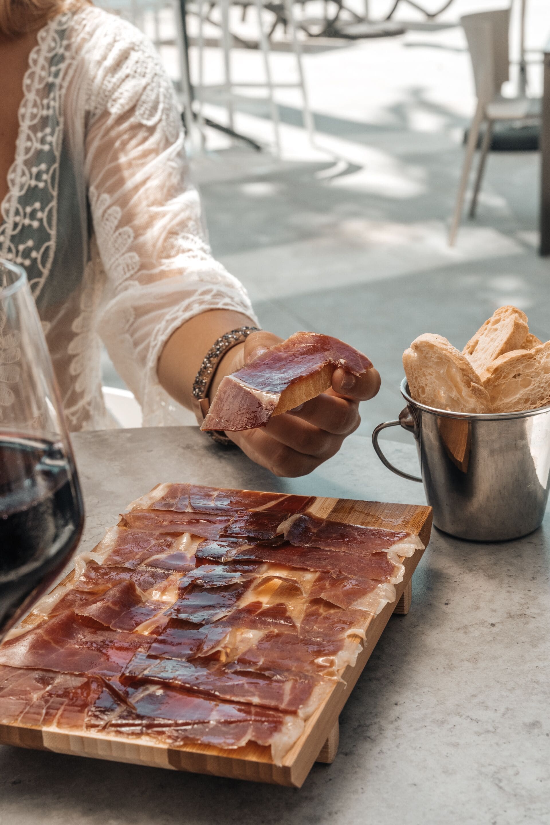 A table with sliced cured meat, a glass of red wine, and a metal cup with bread.