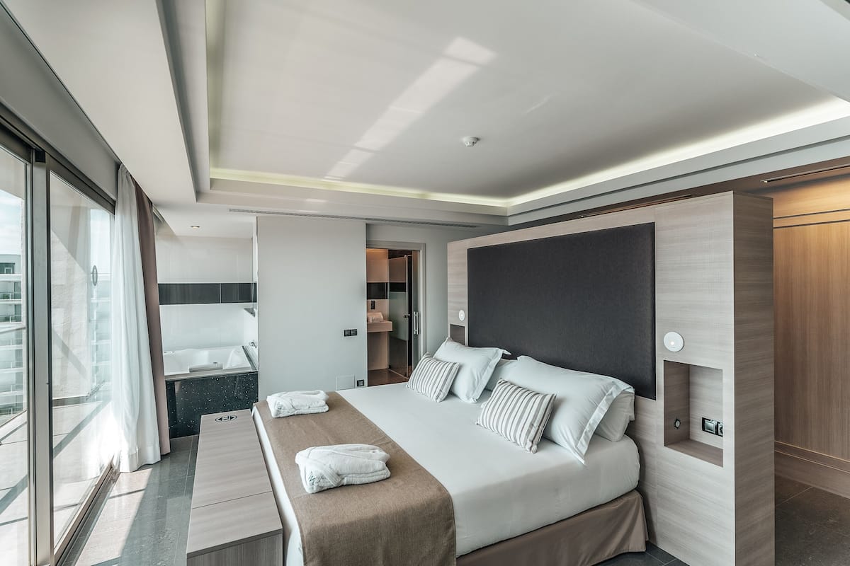 Une chambre d’hôtel moderne dotée d’un grand lit, d’un dressing, d’une salle de bain avec baignoire et offrant une vue par la fenêtre.