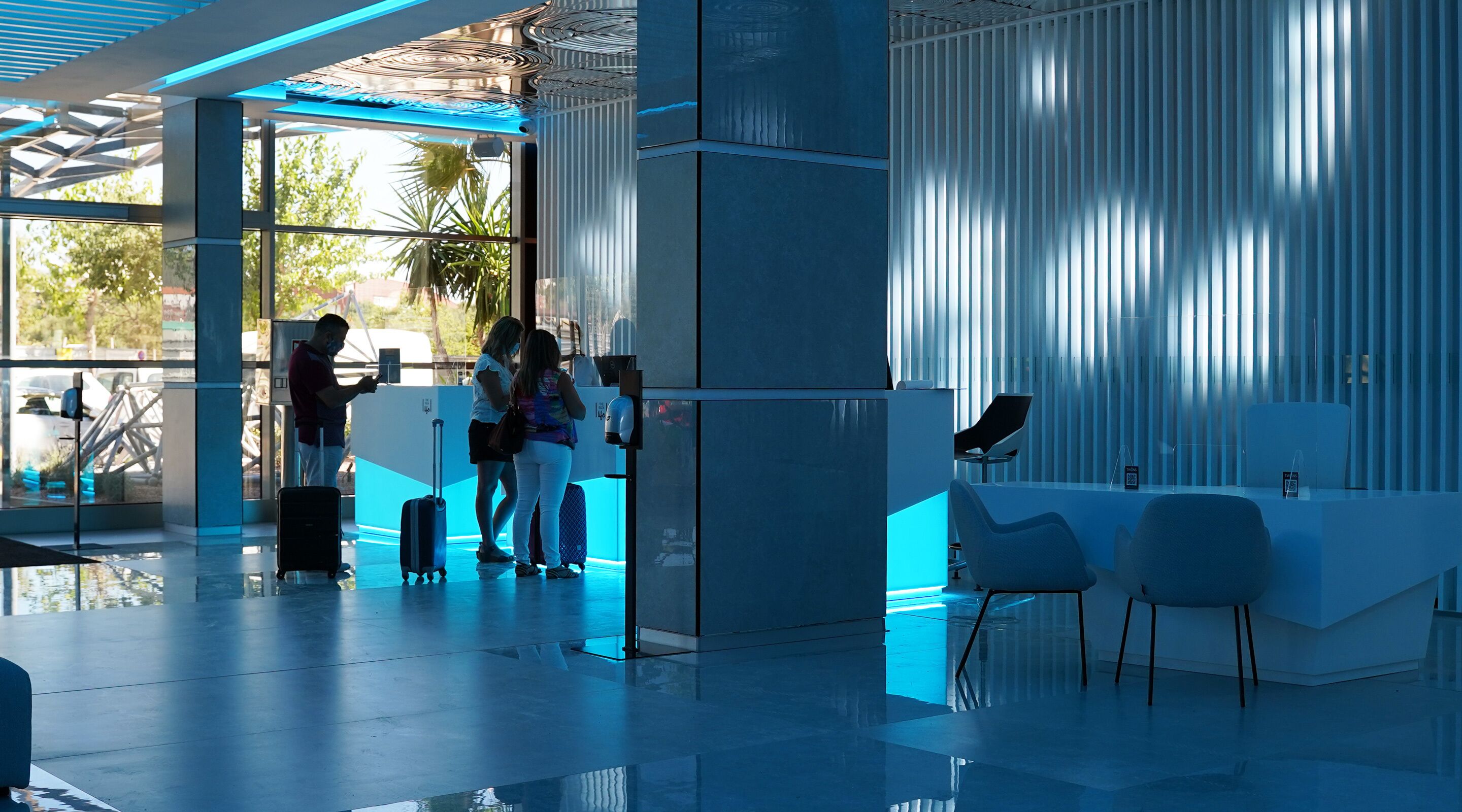 A modern reception area with a glass partition, a seating area with chairs, and a view of palm trees outside.