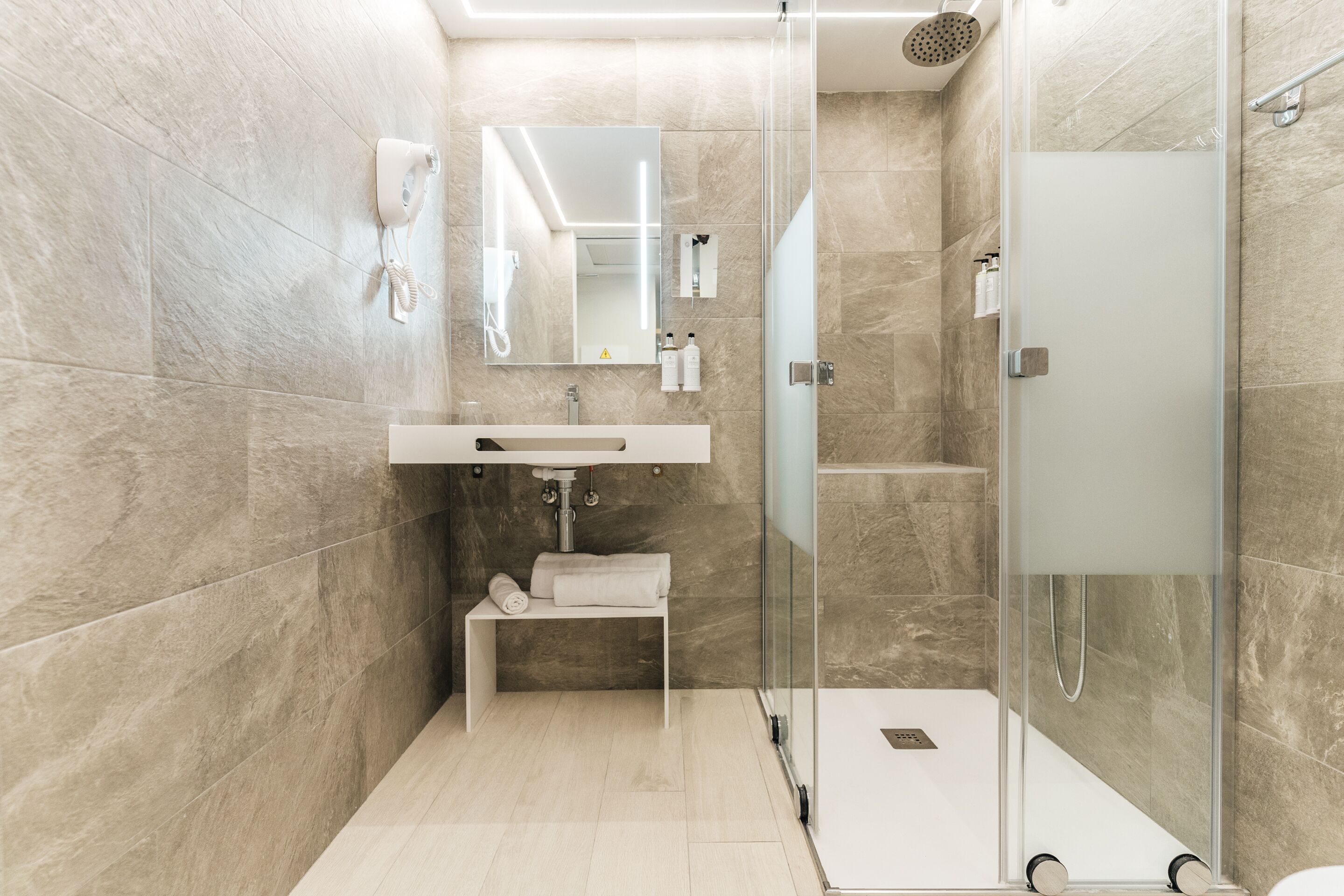 Un baño moderno con ducha acristalada, lavamanos blanco y espejo sobre él.