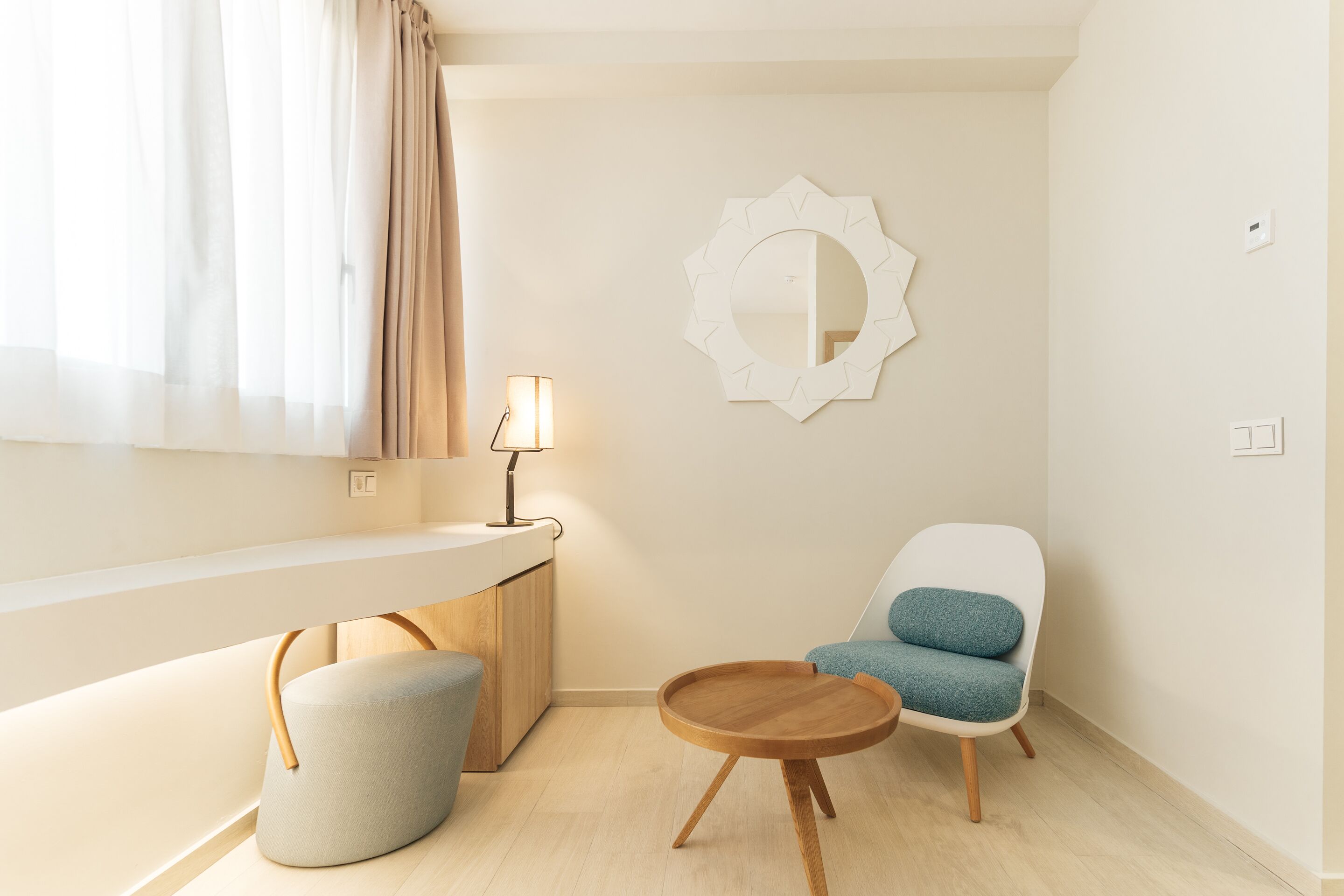 Una habitación moderna con una mesa redonda de madera, una silla blanca con cojín azul, un taburete pequeño y redondo, un espejo montado en la pared y una lámpara de noche.
