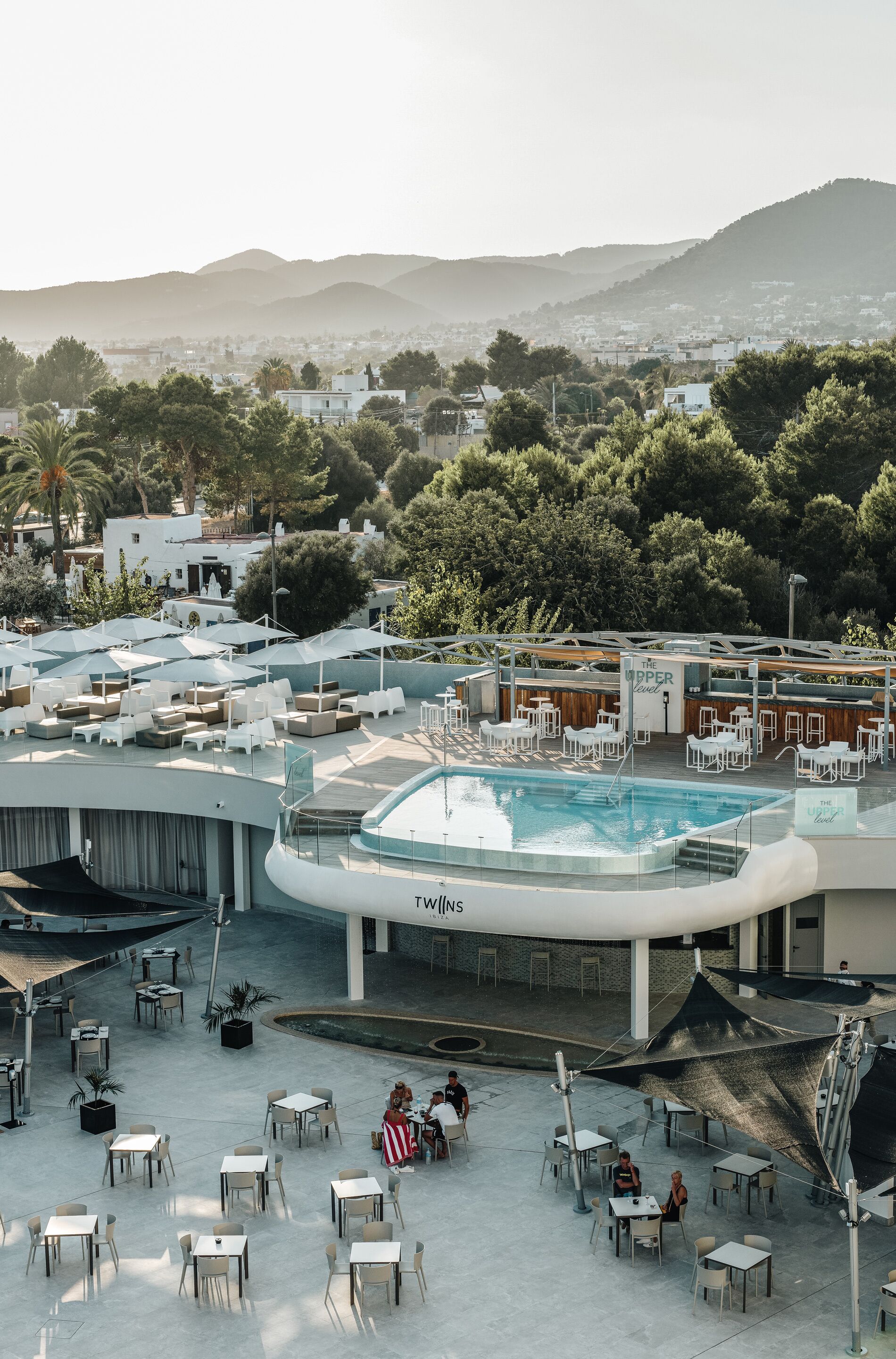 A rooftop pool area with lounge chairs and tables, surrounded by a cityscape and hills.