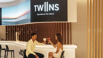 A modern hotel reception area with a large screen displaying the name 'TWILNS' and the tagline 'Experience the island'. There are two bar stools and a counter with a wooden panel design.