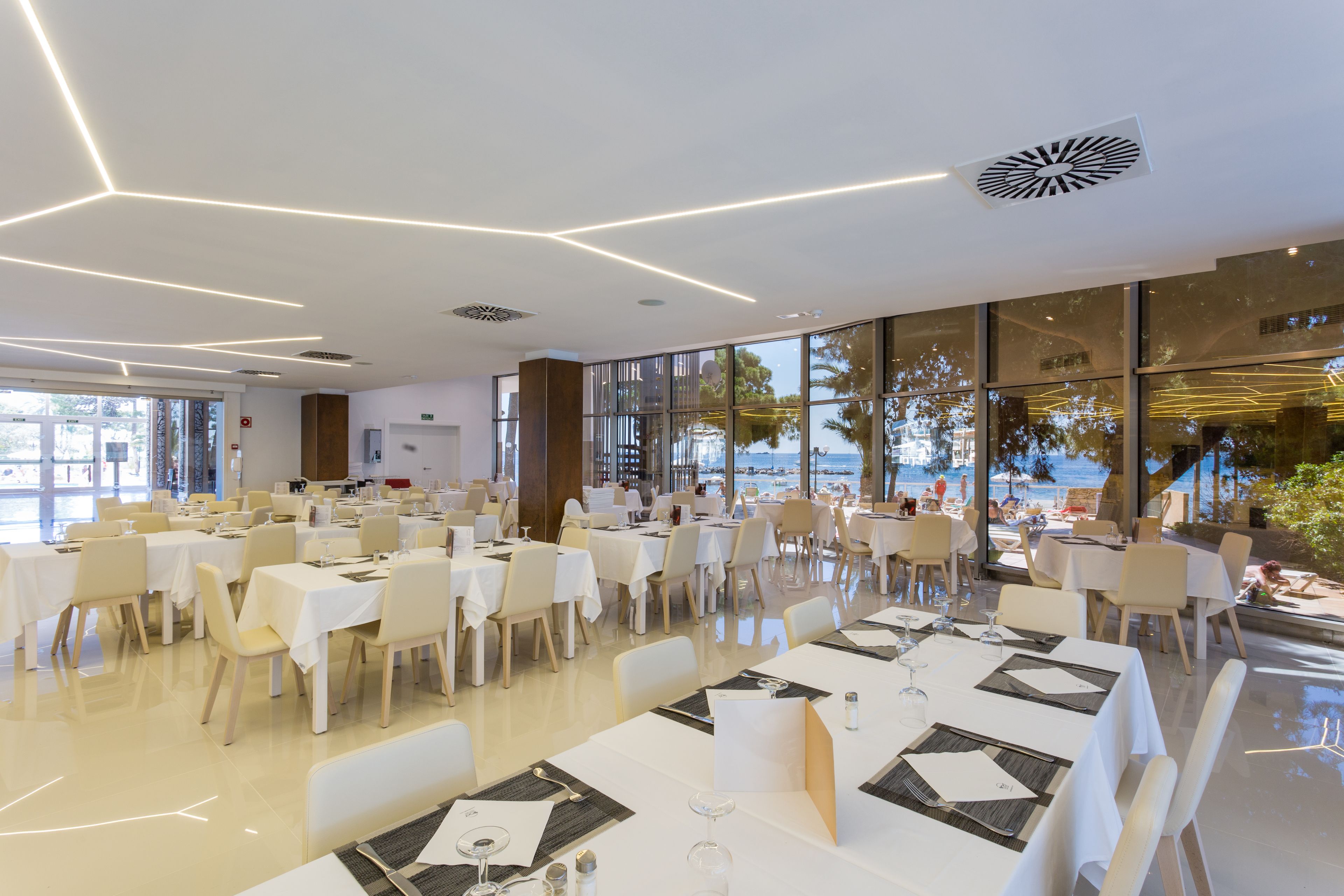 Un amplio comedor con numerosas mesas y sillas, grandes ventanales que ofrecen vistas a la playa y un diseño moderno de techo.