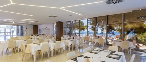 A spacious dining area with multiple tables and chairs, large windows offering a view of the beach, and a modern ceiling design.