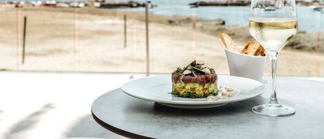 Una mesa con un plato de comida y una copa de vino blanco, frente a una vista de la playa.