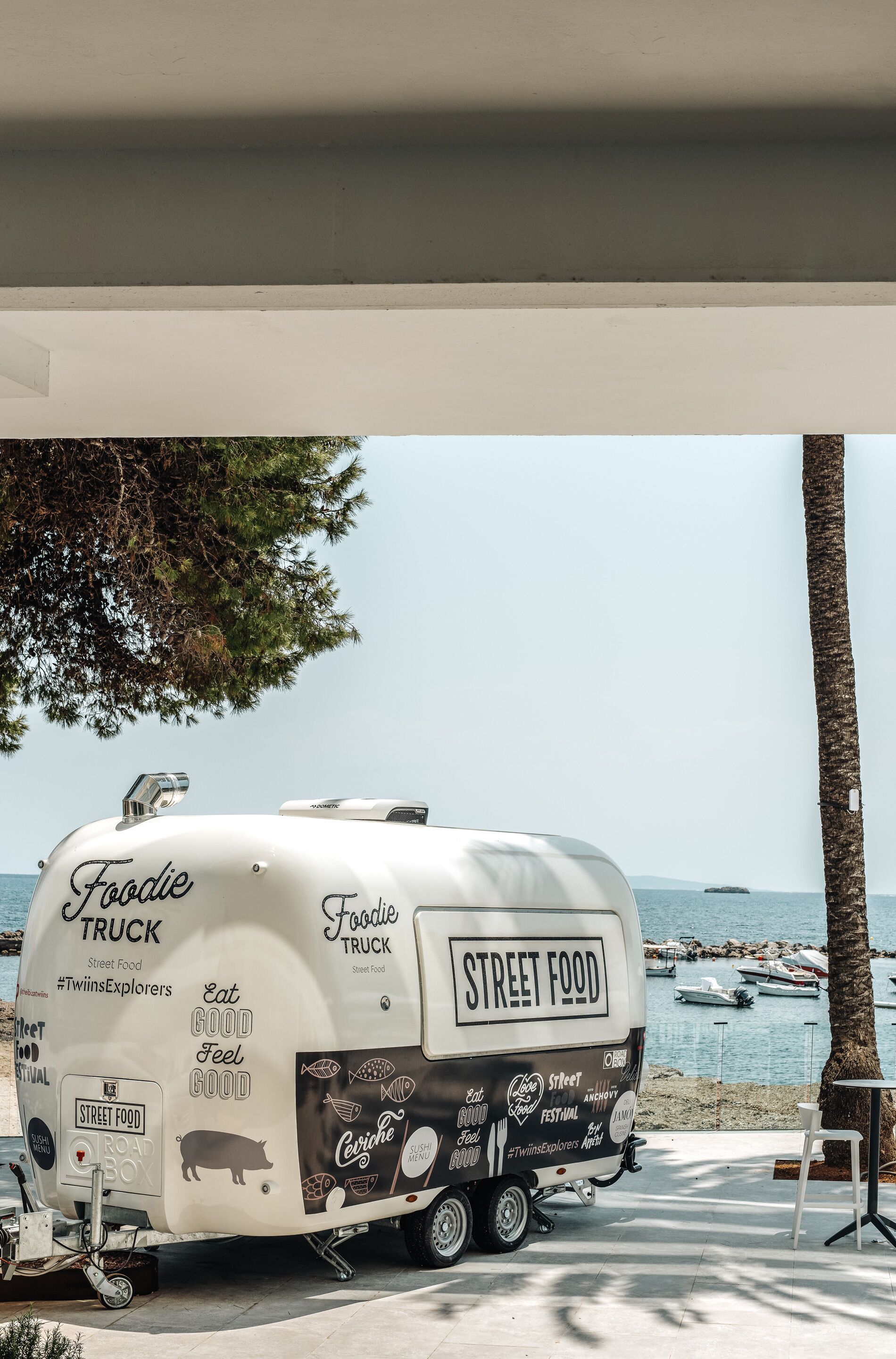 Un camión de comida llamado 'Foodie Truck' estacionado junto a la playa, con vista al mar y barcos al fondo.