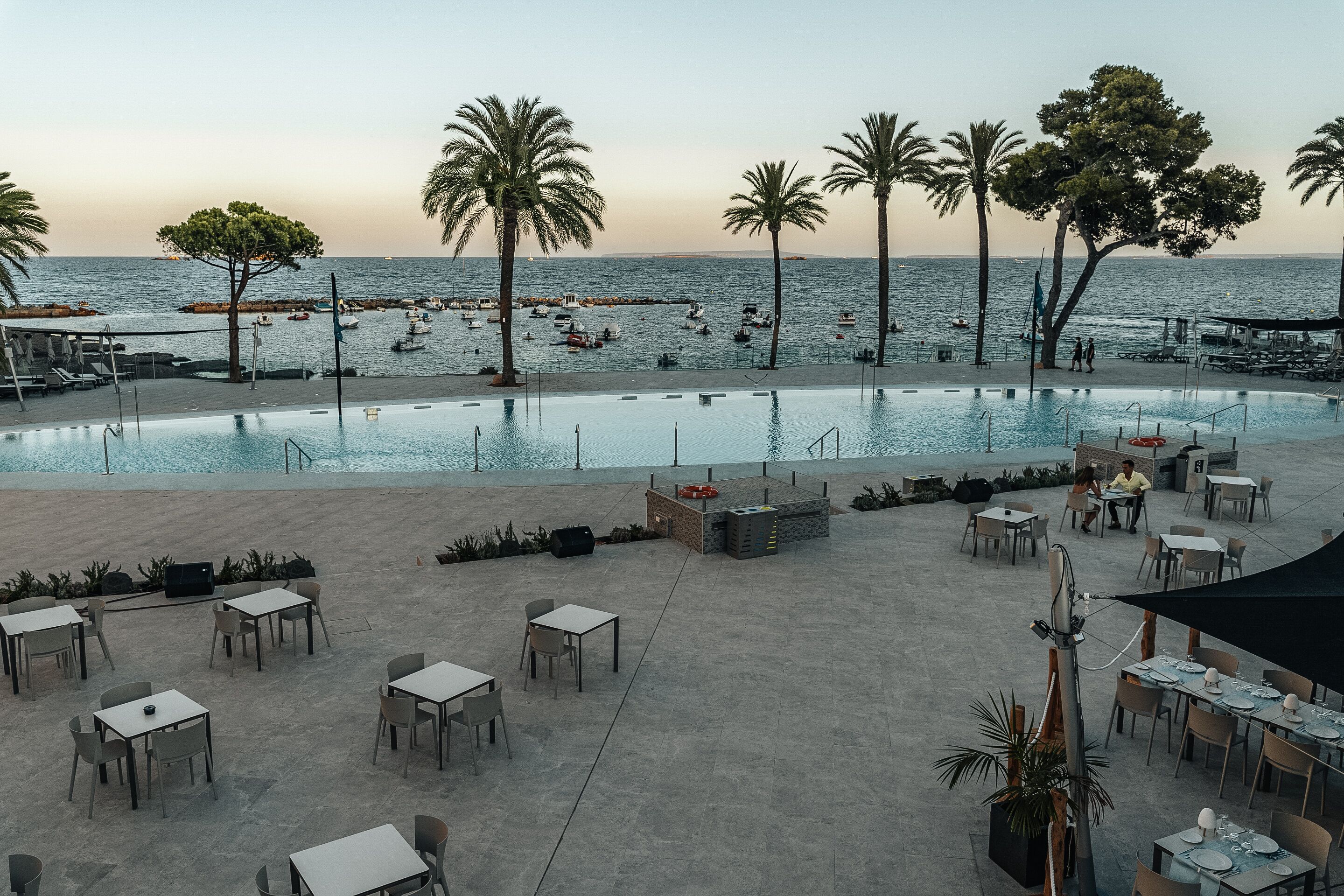 An outdoor pool area with tables and chairs, palm trees, and a view of the sea.