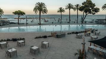 An outdoor pool area with tables and chairs, palm trees, and a view of the sea.