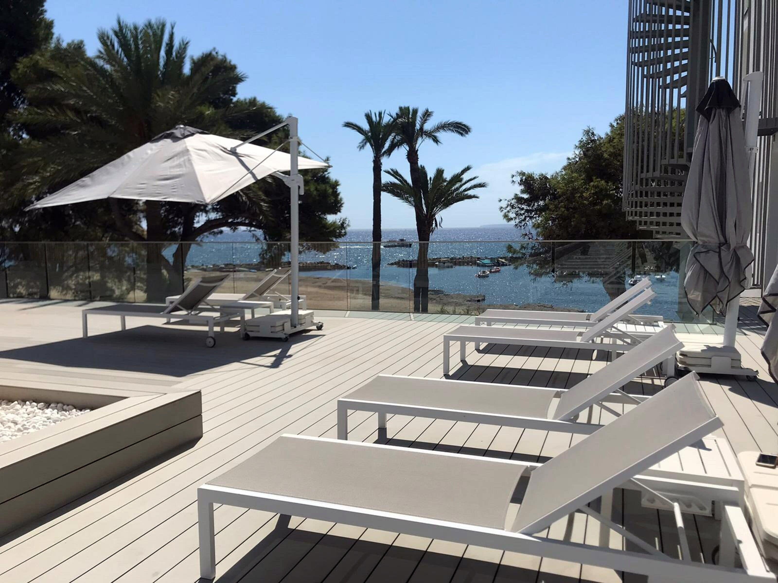 An outdoor deck with white lounge chairs and a large umbrella, overlooking a body of water with boats and palm trees.