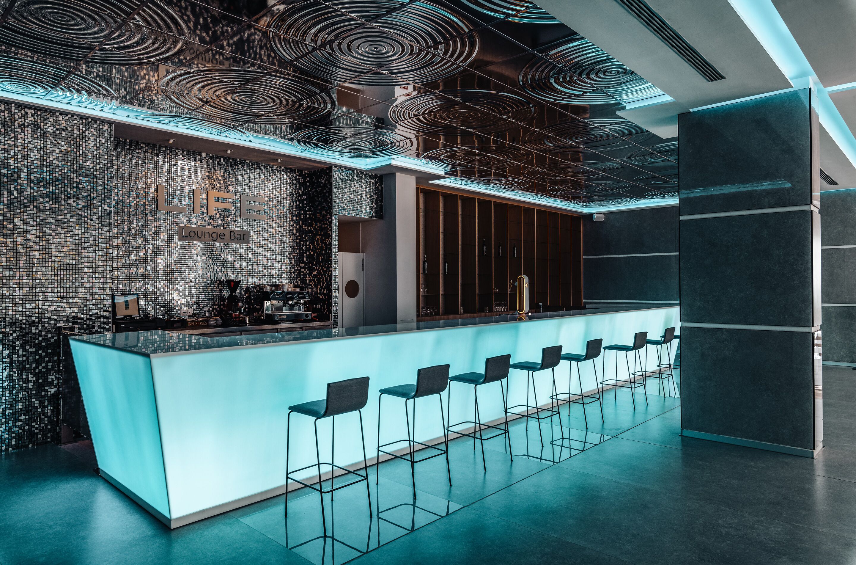 Un bar lounge moderno con una barra central, varios taburetes y un revestimiento mural estampado.