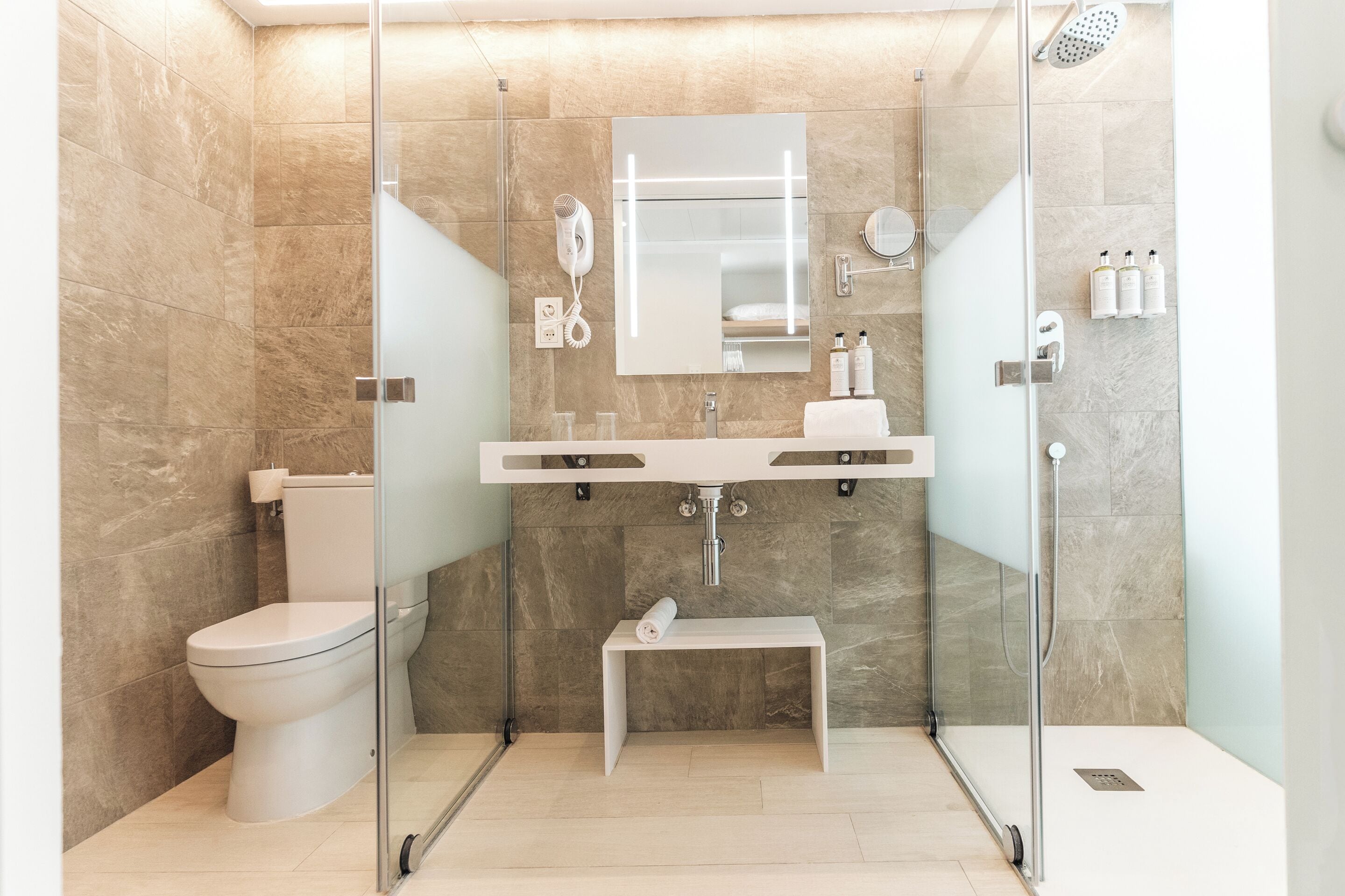 Un baño moderno con ducha de vidrio, inodoro, lavamanos con espejo y un pequeño banco.