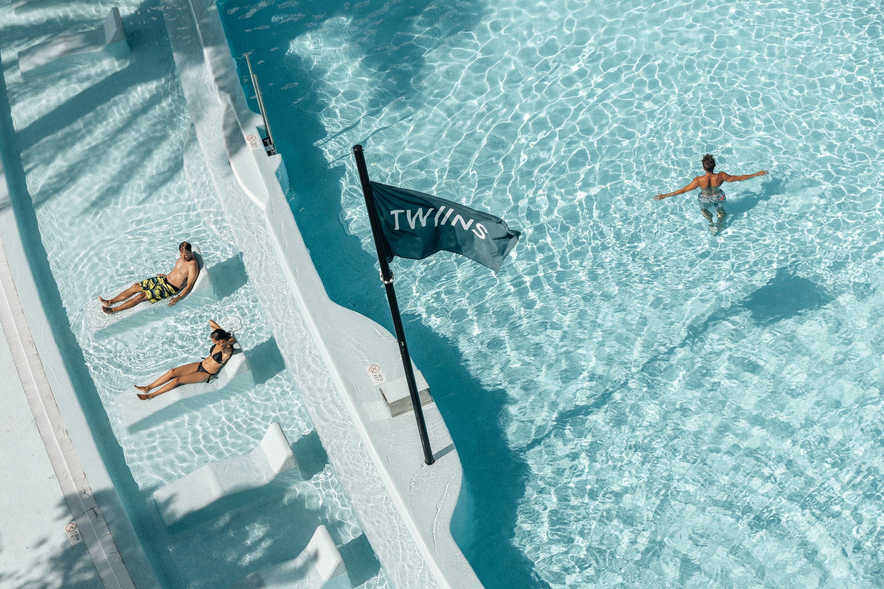 Une vue aérienne d’une piscine à l’eau d’un bleu limpide, avec un drapeau et des personnes se détendant au bord du bassin.