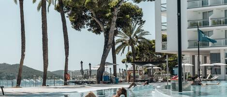 A pool area with lounge chairs and umbrellas, surrounded by palm trees and a building with balconies.
