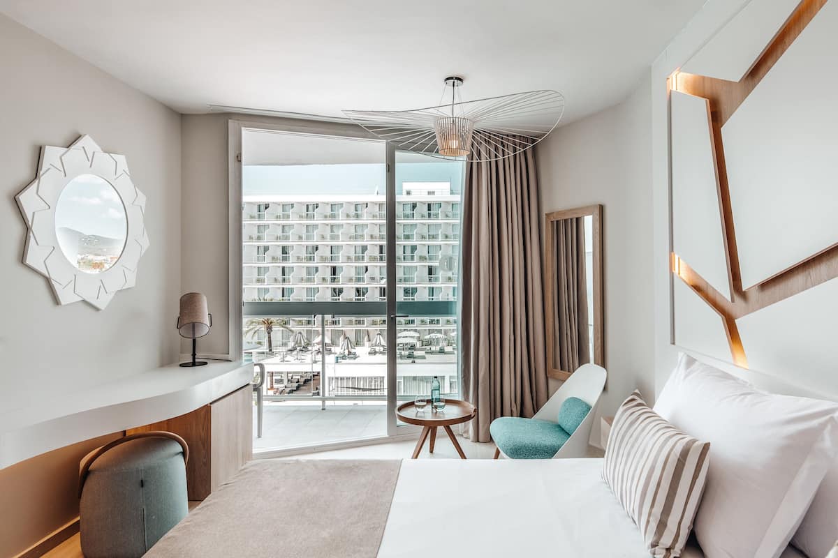 Une chambre d’hôtel avec un grand lit, un coin salon avec une table, une grande fenêtre donnant sur un immeuble et un miroir au mur.