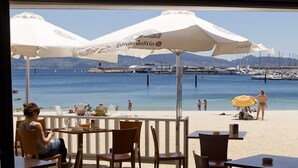 Een restaurant aan de kust met terras, witte parasols en uitzicht op het strand en de jachthaven.