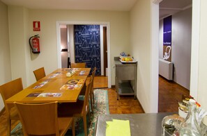 Un comedor con una larga mesa de madera, sillas, un extintor, una puerta con diseño y una pequeña zona de cocina con un refrigerador y una estantería con frutas.