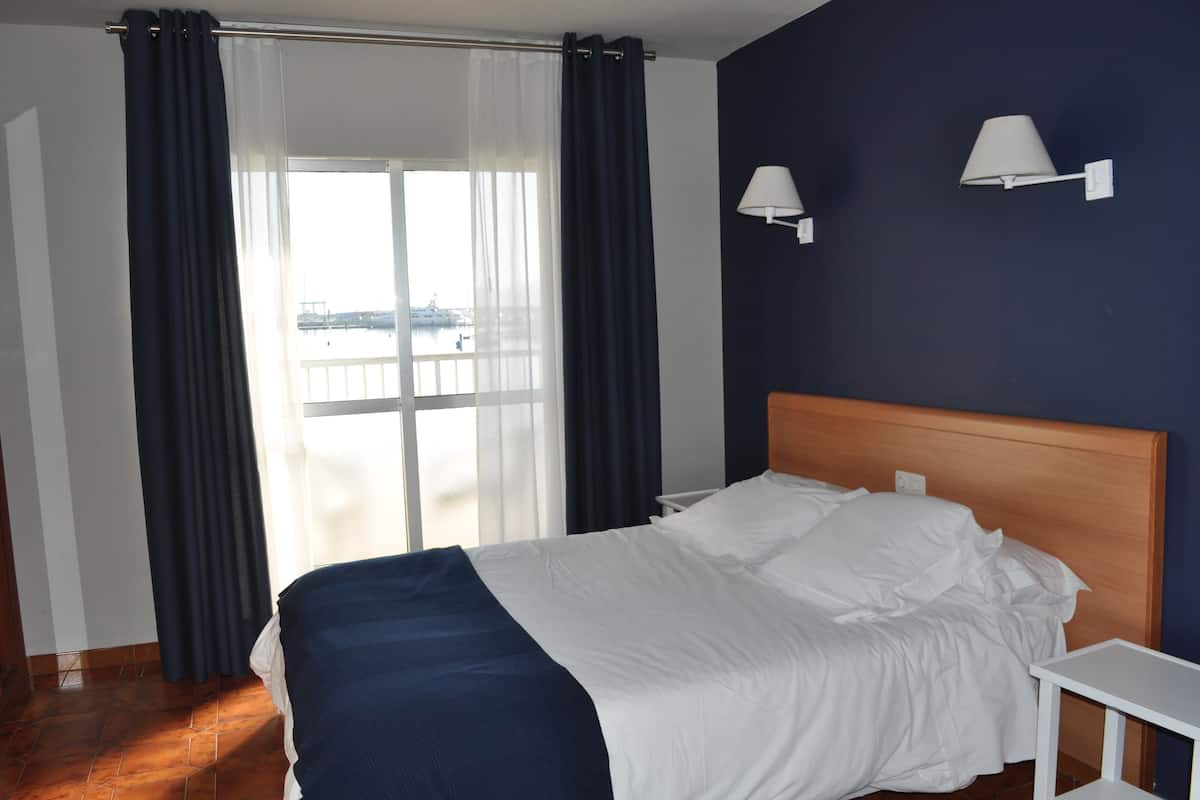 Habitación de hotel con una cama, una mesita de noche, dos lámparas de pared y vista a una masa de agua a través de un ventanal con cortinas.