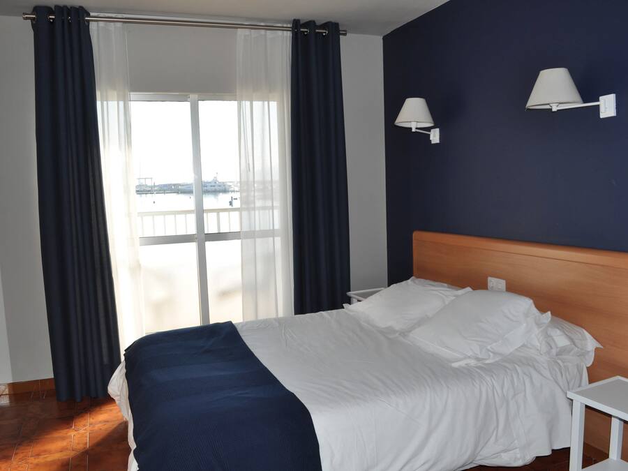 A hotel room with a bed, a nightstand, two wall-mounted lamps, and a view of a body of water through a large window with curtains.