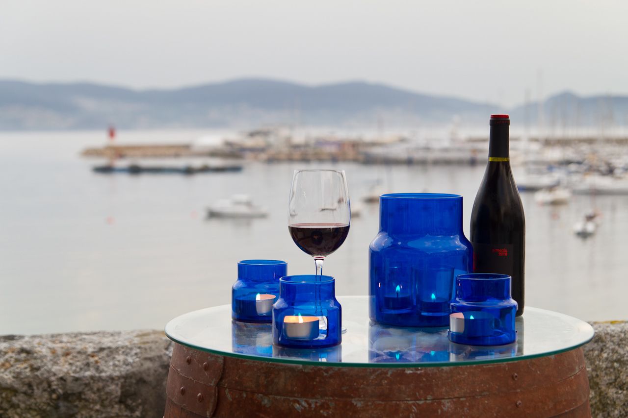 A glass table with a wine glass, a bottle, and blue candle holders by a waterfront.