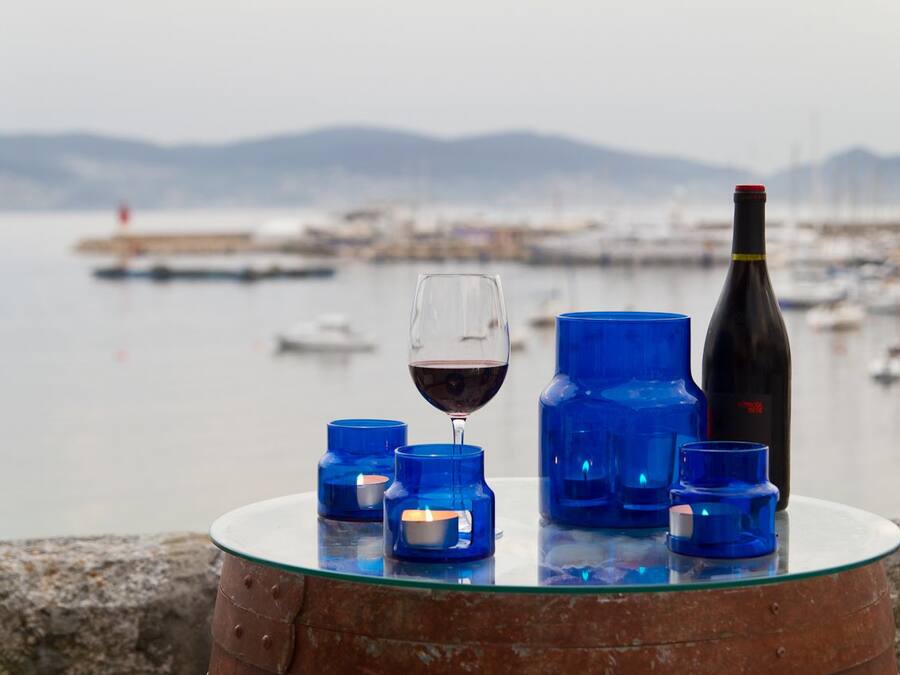 A glass table with a wine glass, a bottle, and blue candle holders by a waterfront.