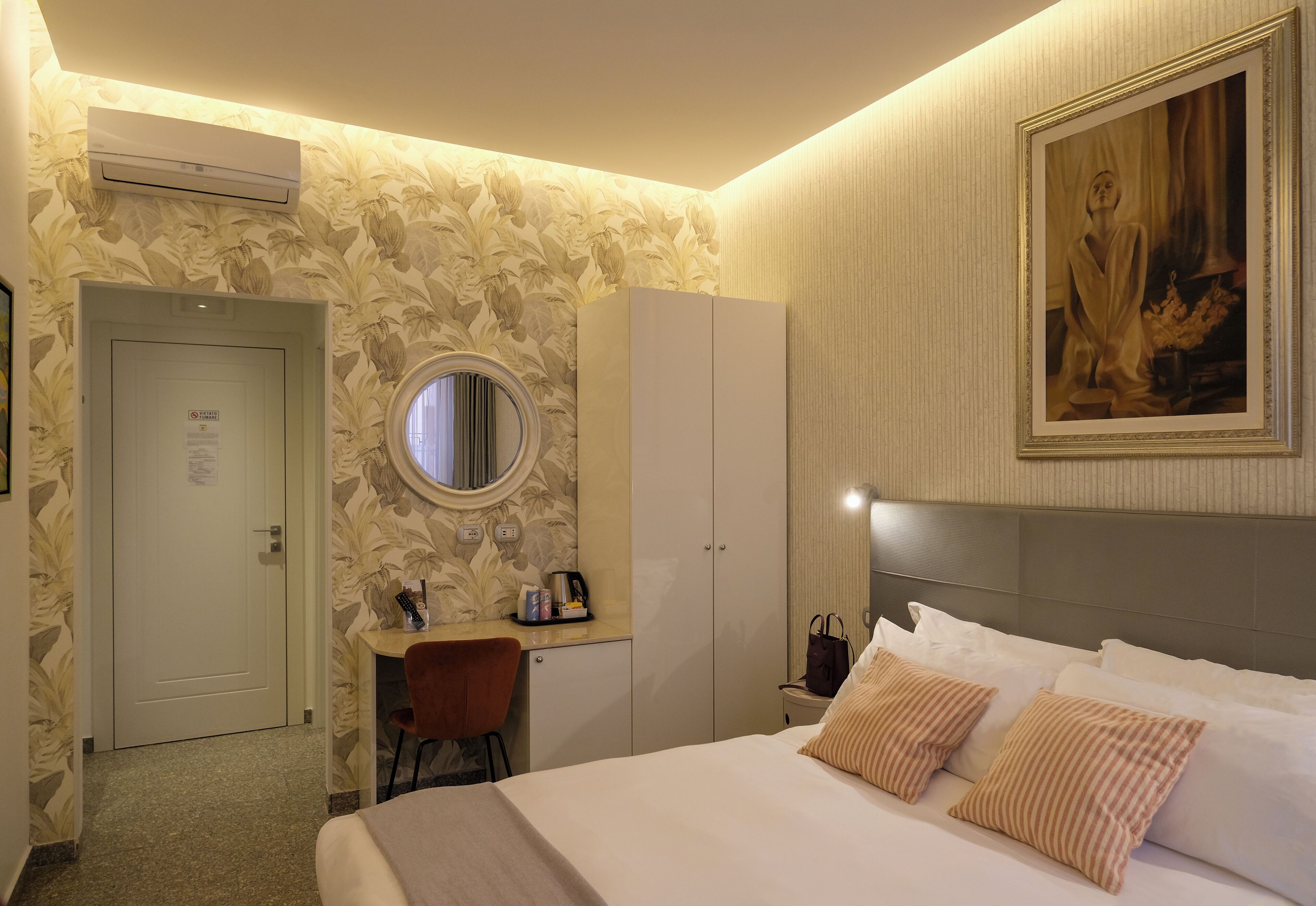 A hotel room with a bed, a desk, a chair, a wardrobe, an air conditioning unit, a painting, and a door with a sign.
