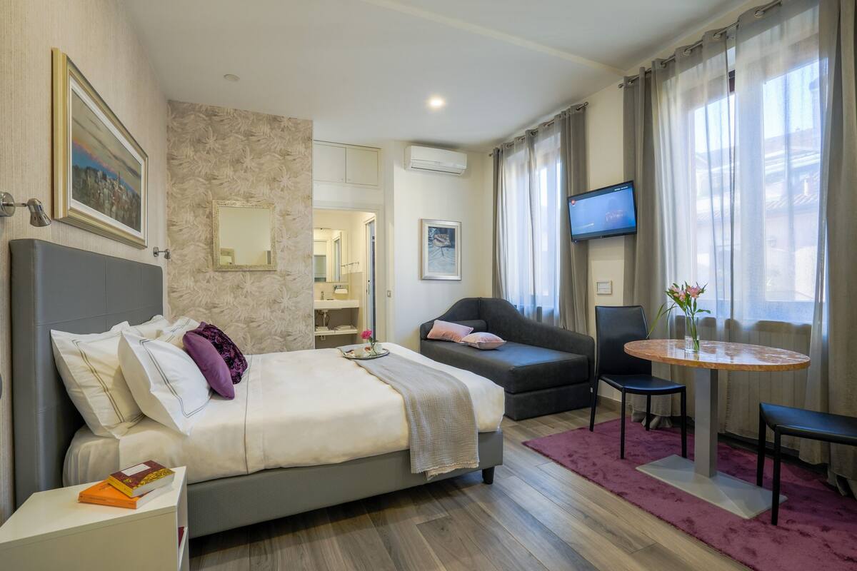 A hotel room with a bed, a sofa, a TV, and a small table with a vase of flowers.