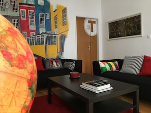 Ein Wohnzimmer mit einem schwarzen Sofa, einem Couchtisch mit Büchern, einem Globus, einem Wandbild mit bunten Gebäuden und einer Tür mit der Beschriftung "01".