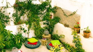 A small, enclosed outdoor space with a white wall, a wooden table, two round seating areas with cushions, and various potted plants. - Terrace Lisbon Hostel (Lisbon)