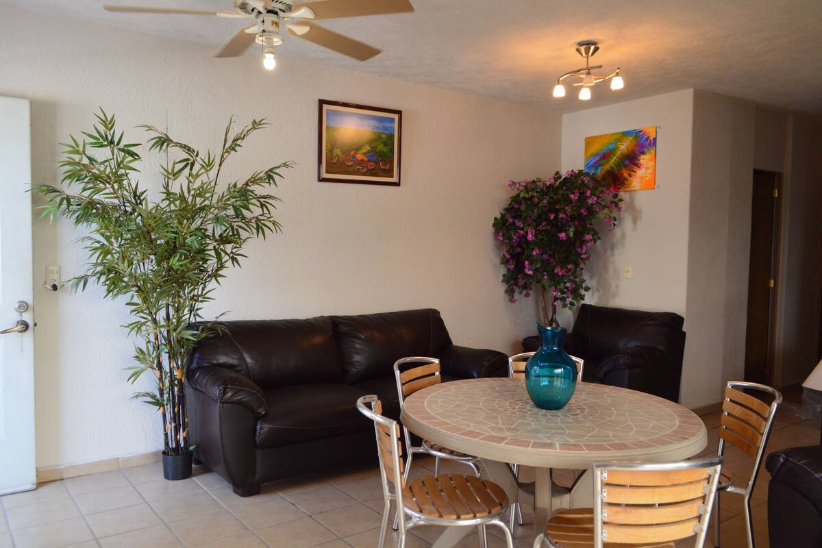 A living room with a brown leather sofa, a round table with chairs, a ceiling fan, and a potted plant.