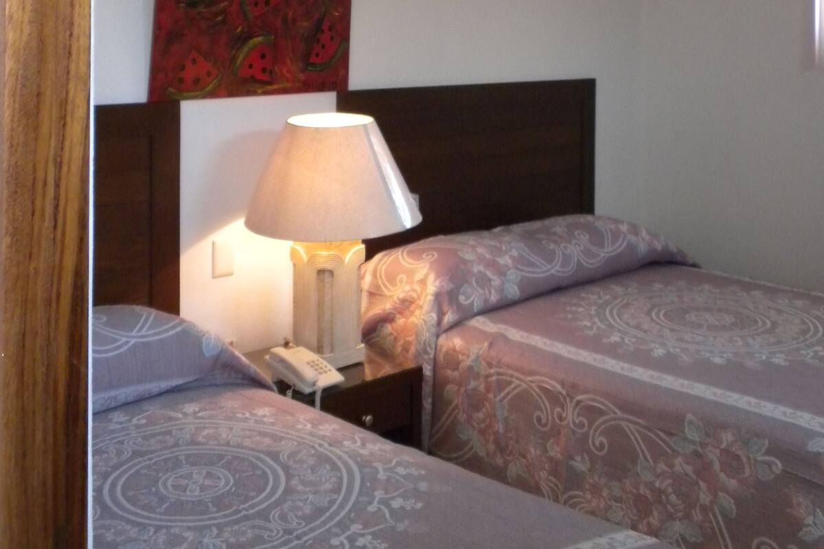 A hotel room with two beds, a nightstand with a lamp, a telephone, and a ceiling fan.
