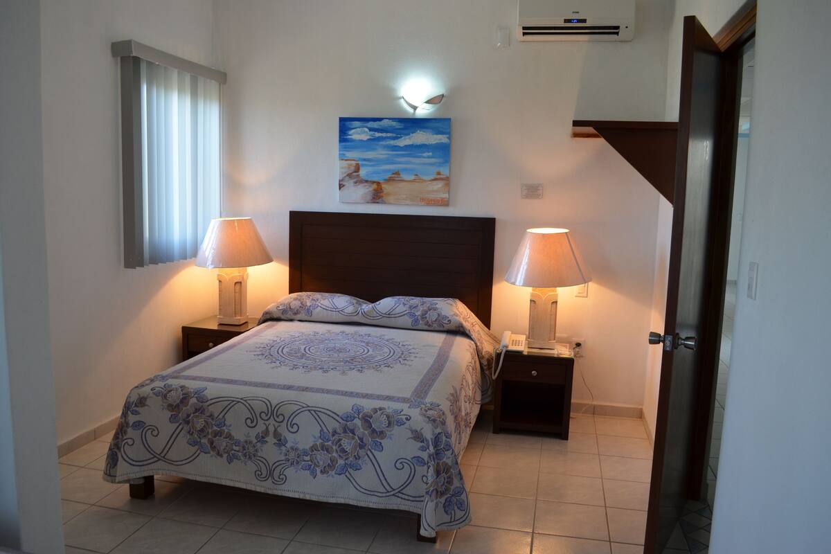A bedroom with a bed, two bedside tables with lamps, a painting on the wall, and a door.