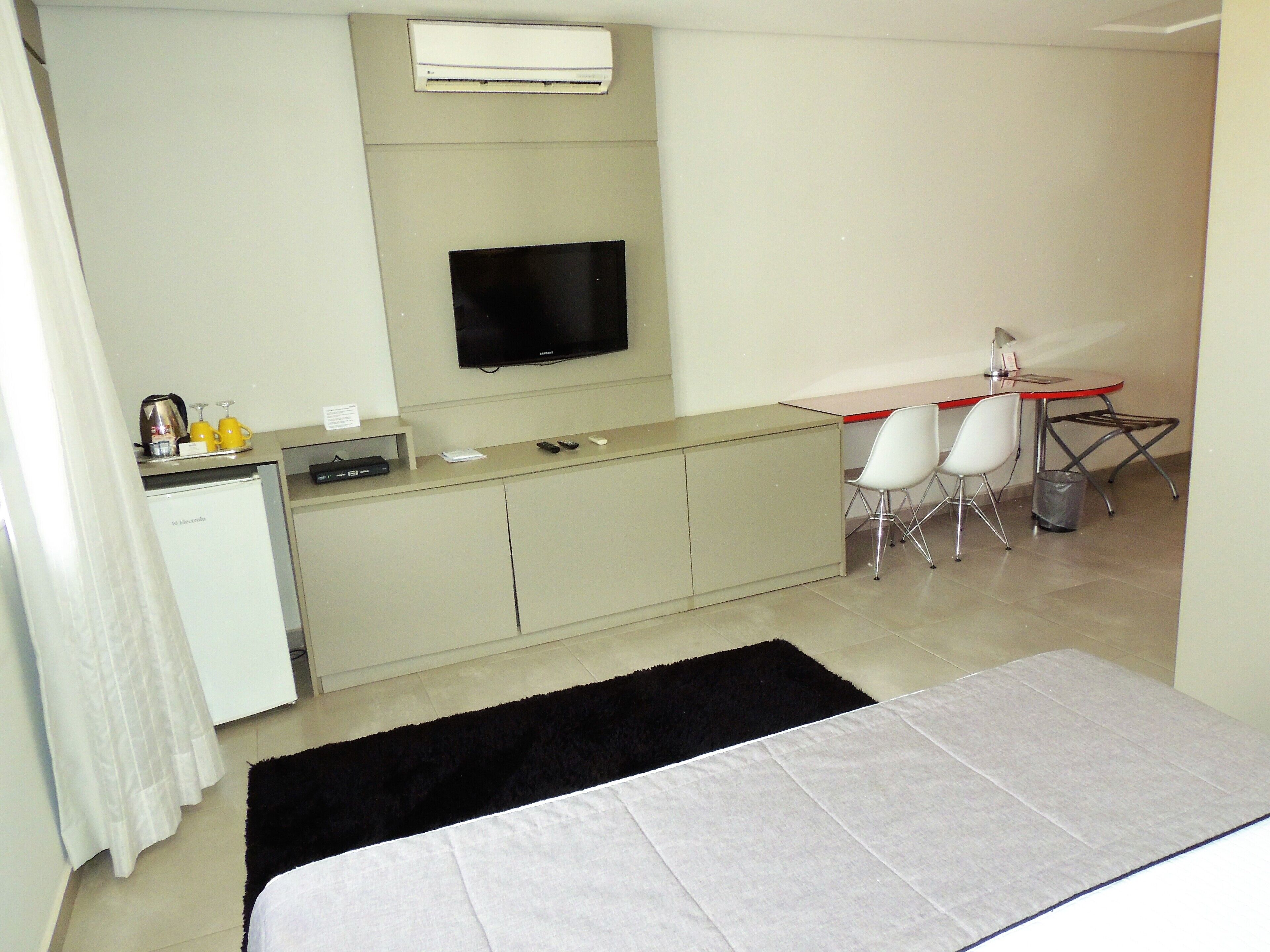 Quarto de hotel pequeno com cozinha compacta, televisão de tela plana, cama e escrivaninha com duas cadeiras.