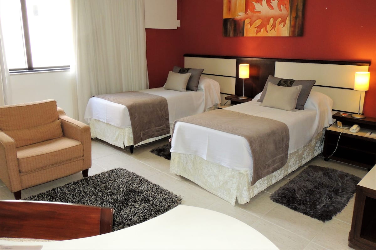 Habitación de hotel con dos camas, una silla, un escritorio, una lámpara y un cuadro en la pared.