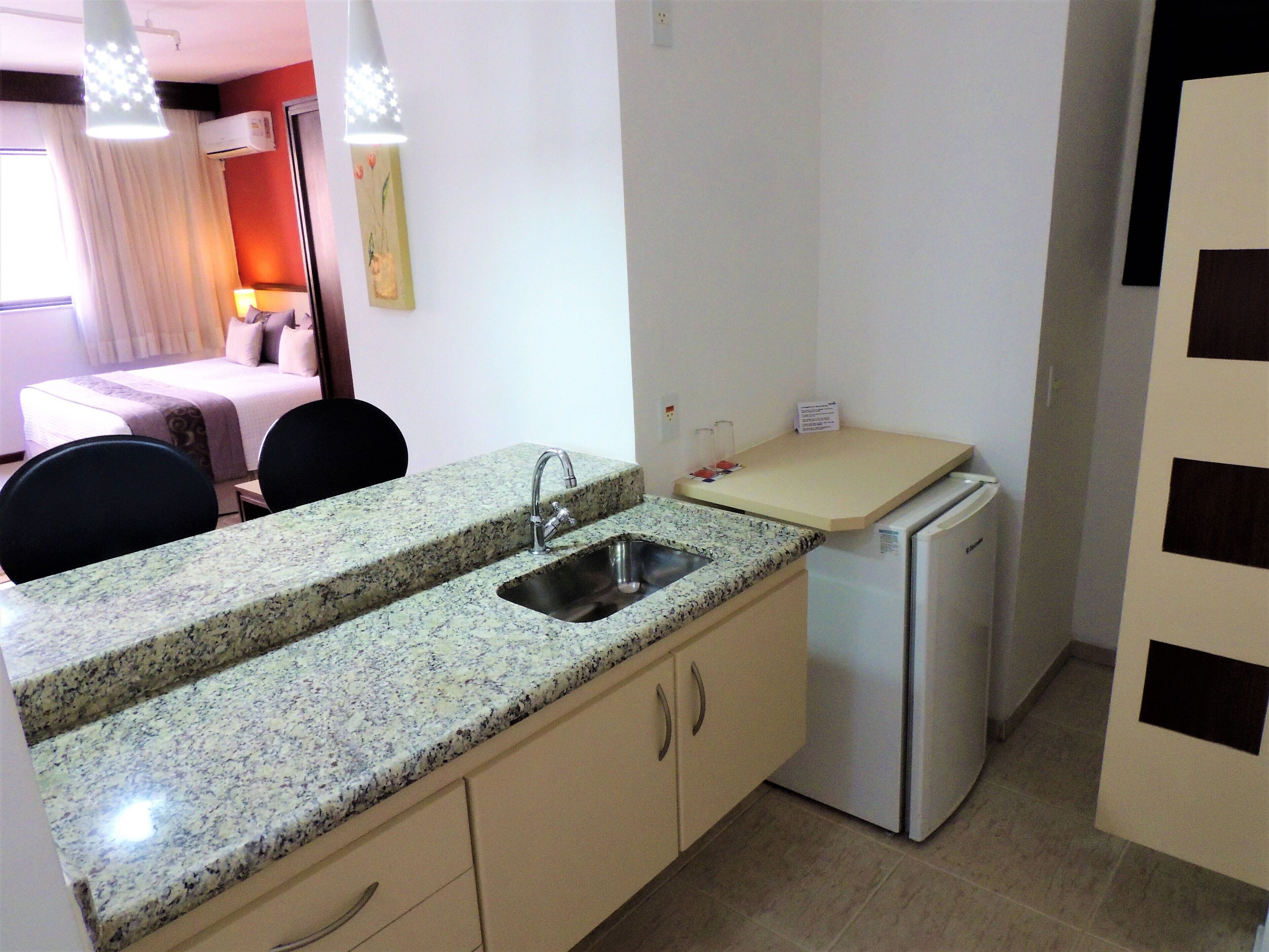 Una cocina compacta con encimera de granito, un lavamanos, un refrigerador y una zona de comedor con una cama al fondo.