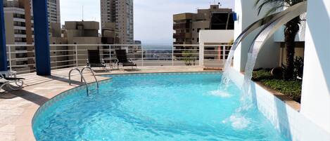 A rooftop pool with clear blue water, a water slide, and a view of the city skyline.