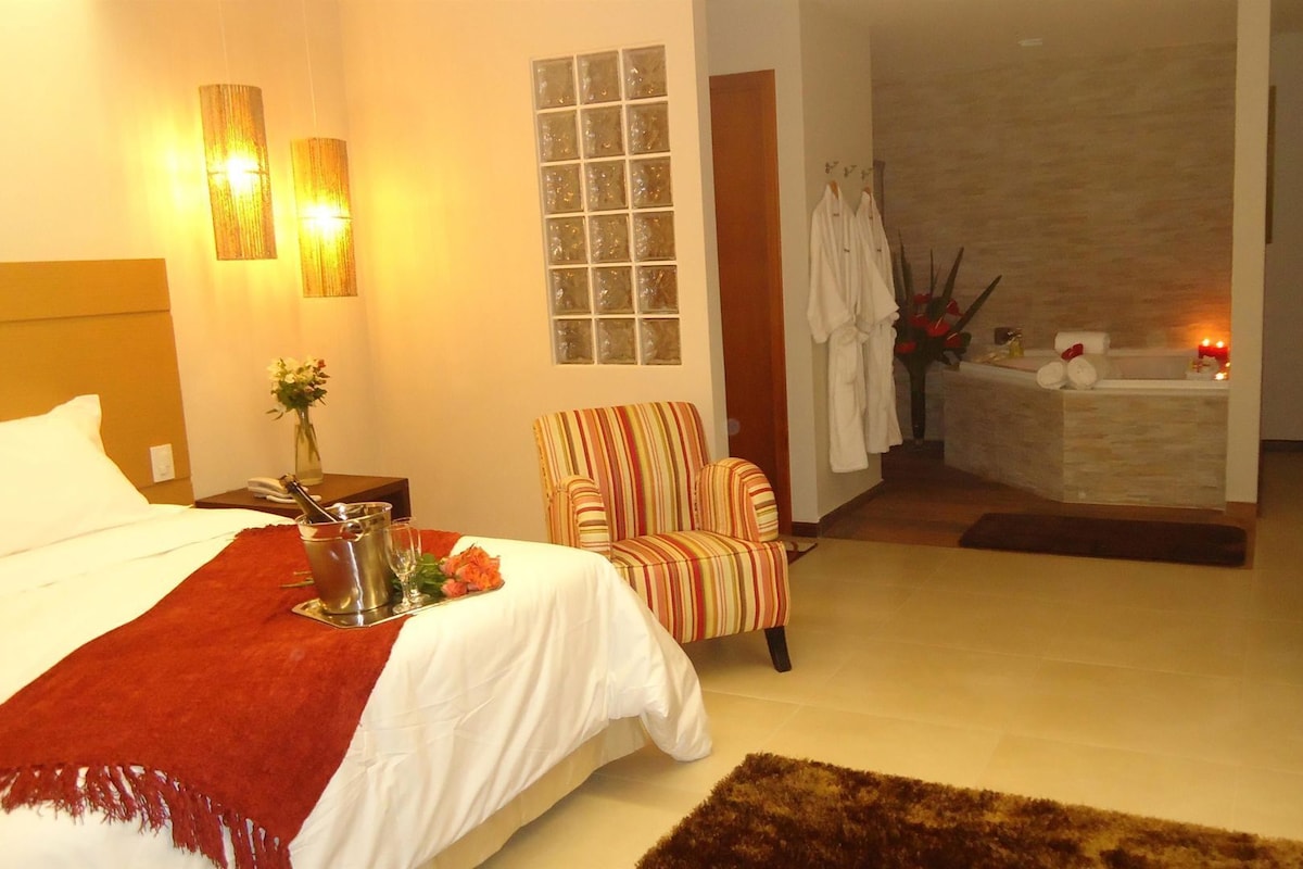 Une chambre d’hôtel avec un lit, un fauteuil à rayures, une petite table avec un vase, une baignoire et une autre petite table avec un vase.
