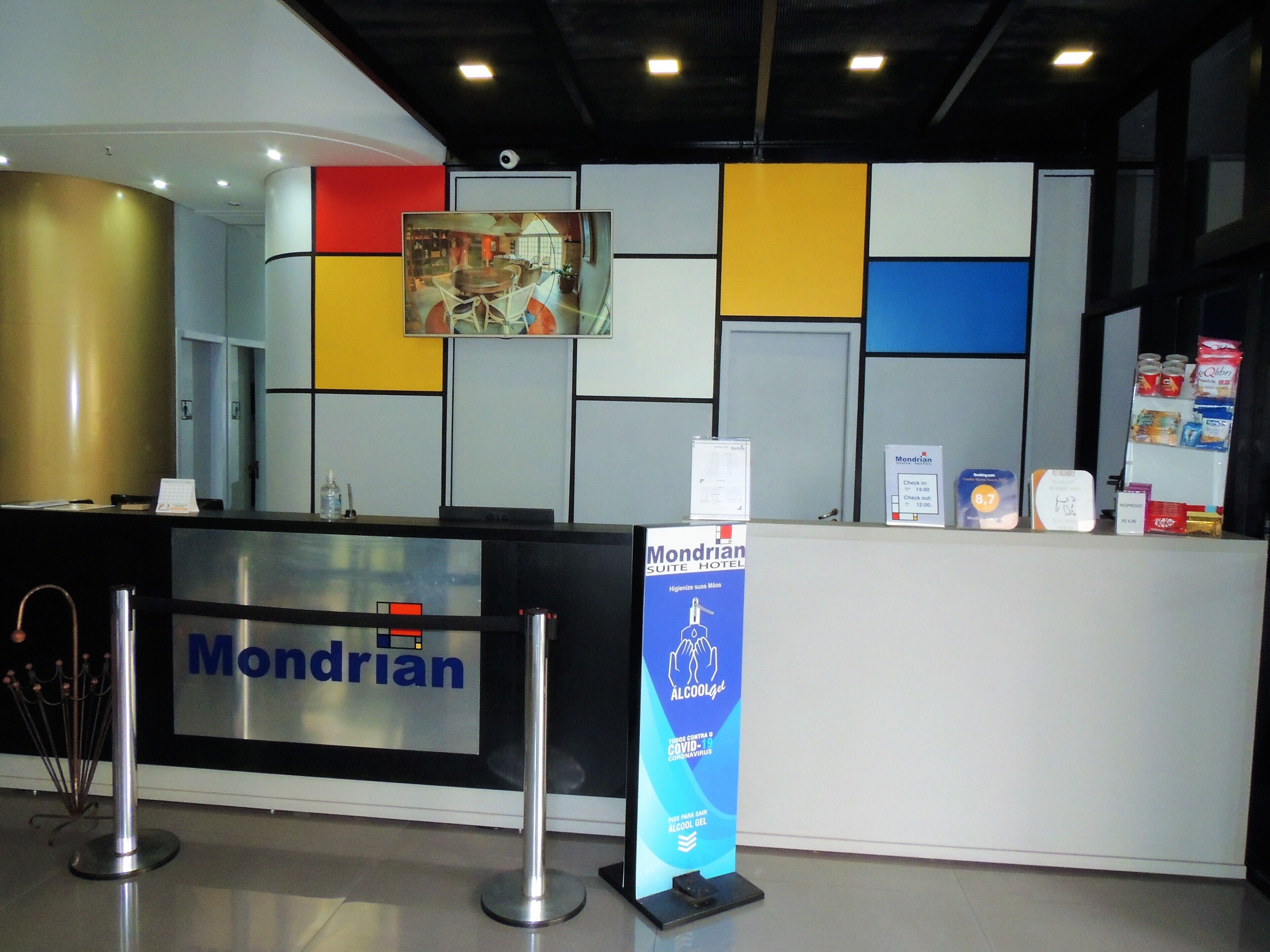 Uma mesa de recepção de hotel com uma placa 'Mondrian', um dispensador de álcool em gel e uma placa de identificação 'Mondrian Hotel'.