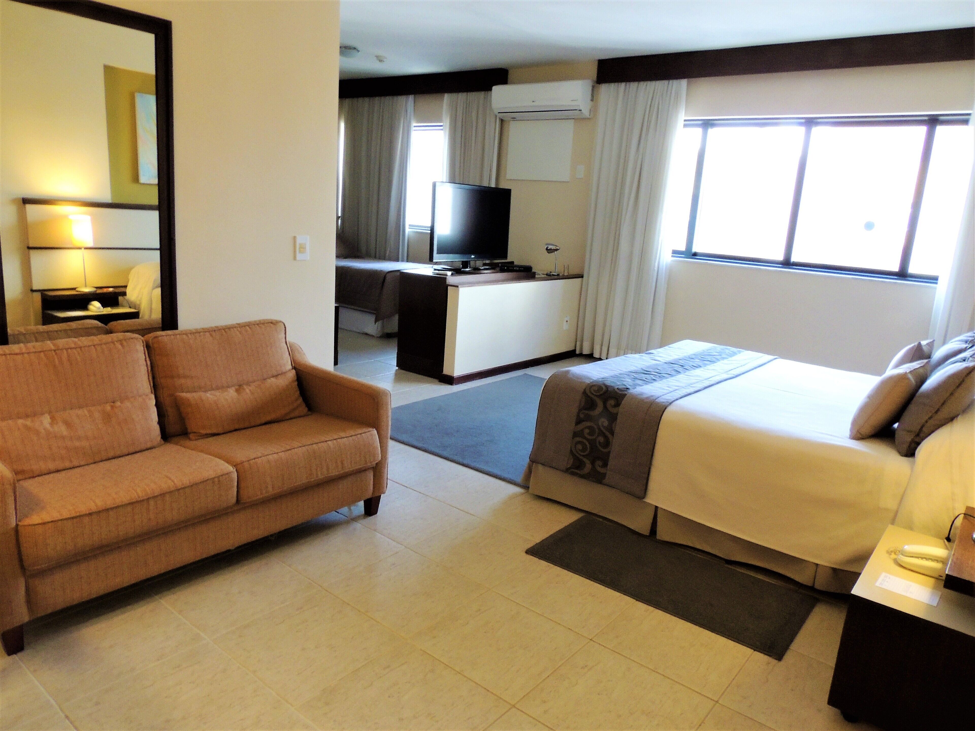 Una habitación de hotel con sofá, cama, escritorio con televisor y ventana con cortinas.