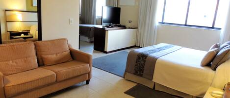 Una habitación de hotel con sofá, cama, escritorio con televisor y ventana con cortinas.
